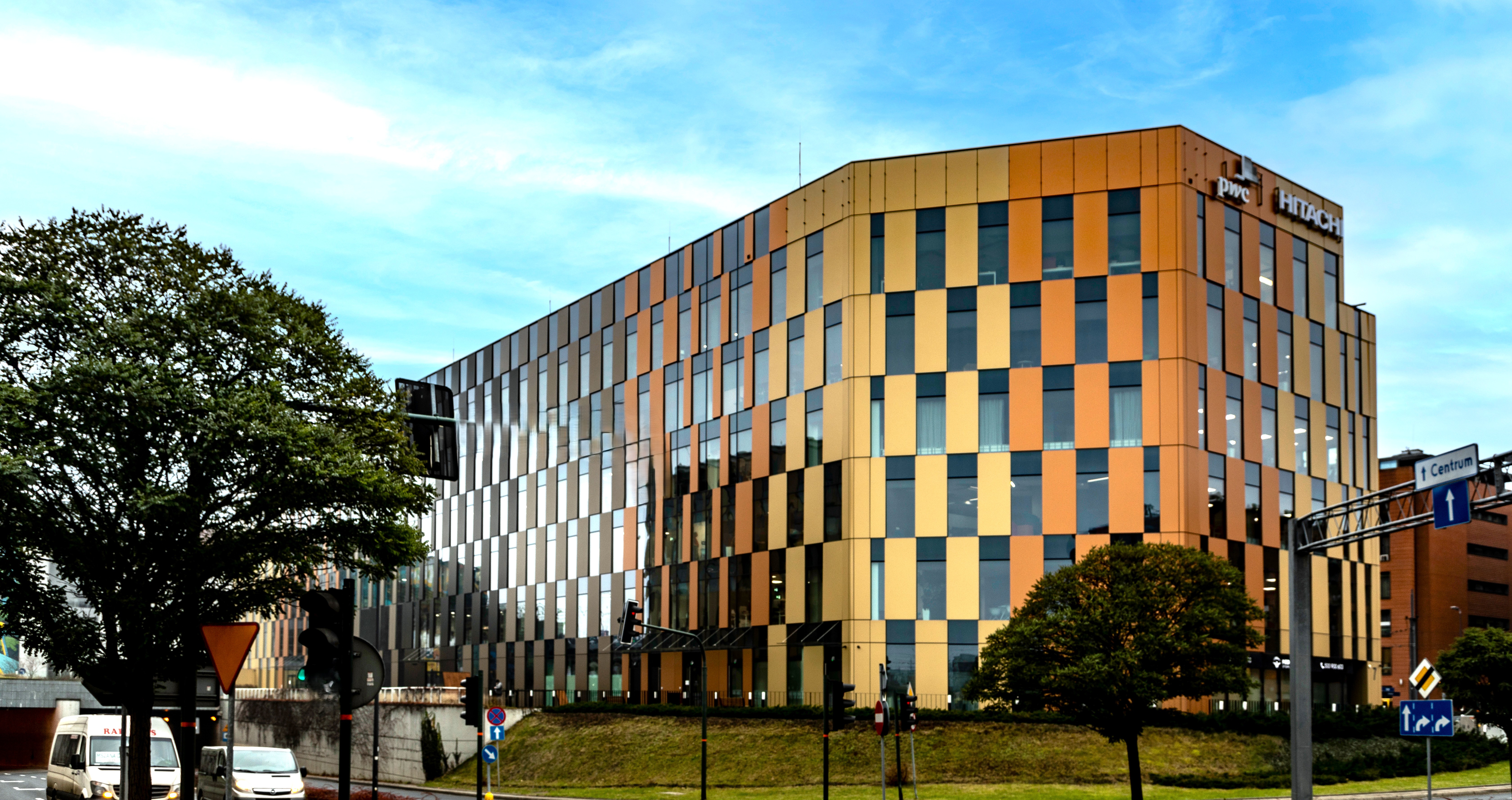 A modern office building with a golden and glass facade, featuring vertical panels of varying shades. The building is surrounded by trees and urban infrastructure, with a bright blue sky in the background