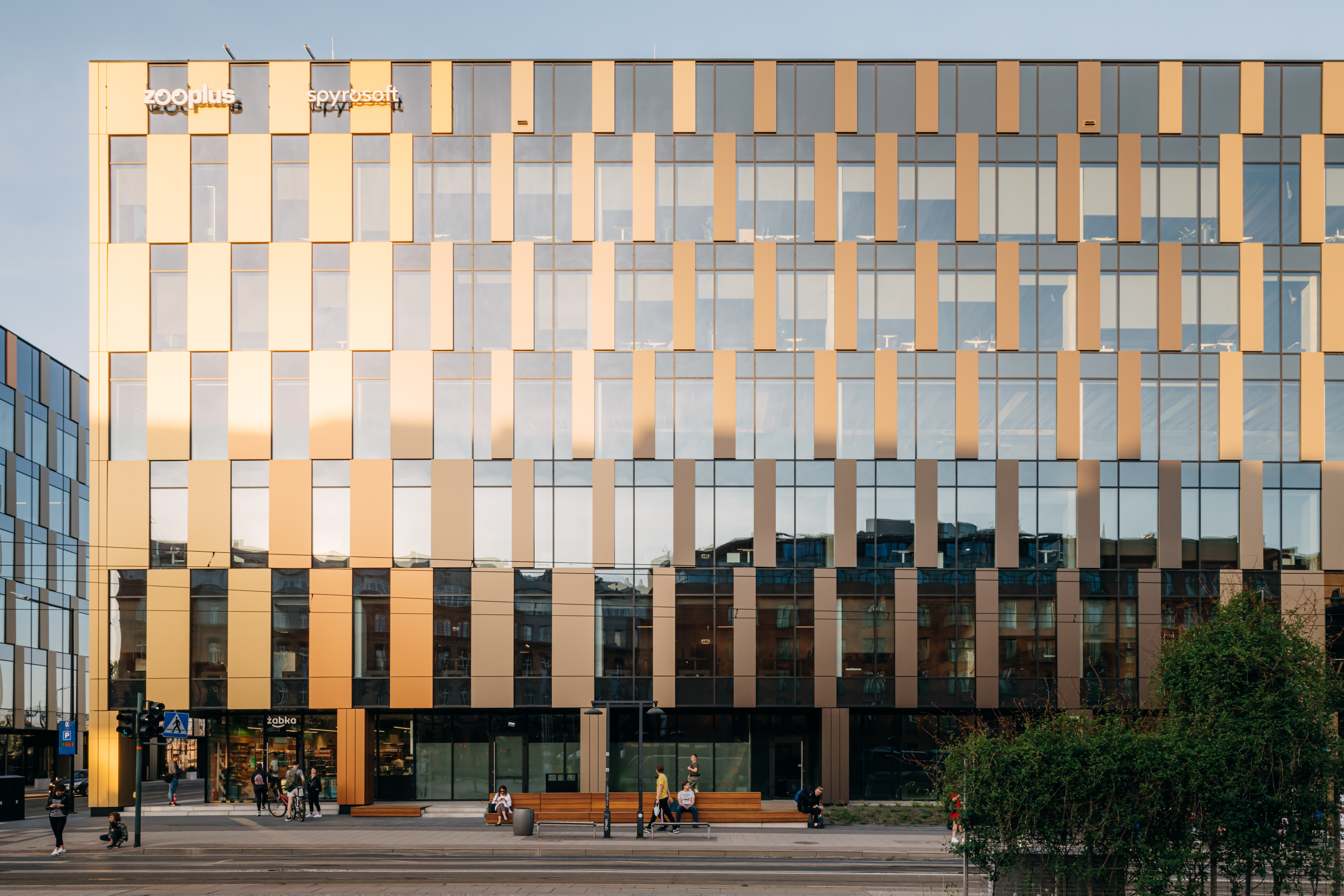 A modern rectangular building with a golden and glass facade, composed of vertical panels in alternating tones. The building faces a street with pedestrians walking along the sidewalk and a few trees in the foreground.