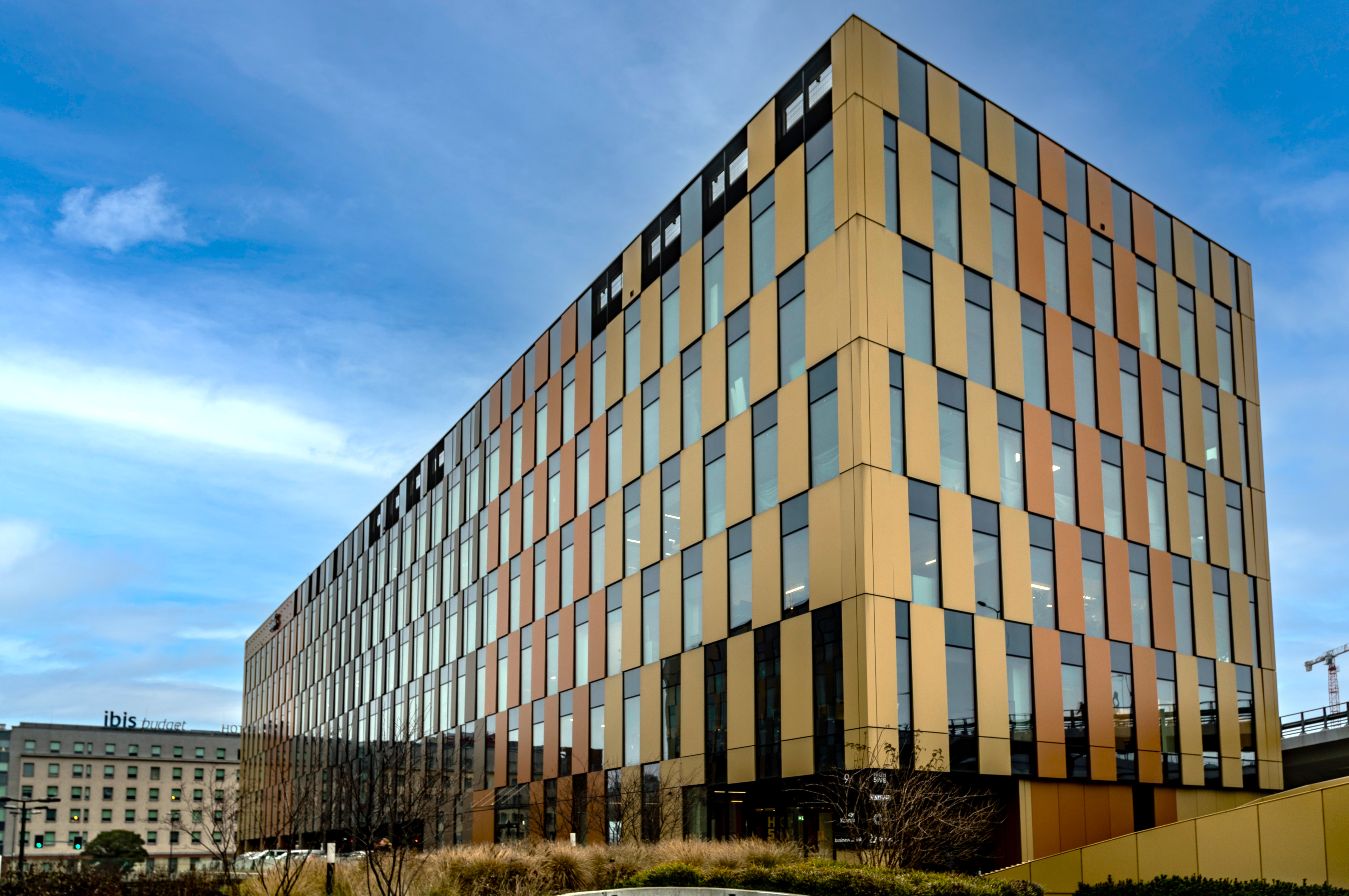 A modern, angular office building with a golden, bronze, and glass facade made of vertical panels. The structure stands under a partly cloudy blue sky, with landscaped grass and plants in the foreground and city buildings in the background.