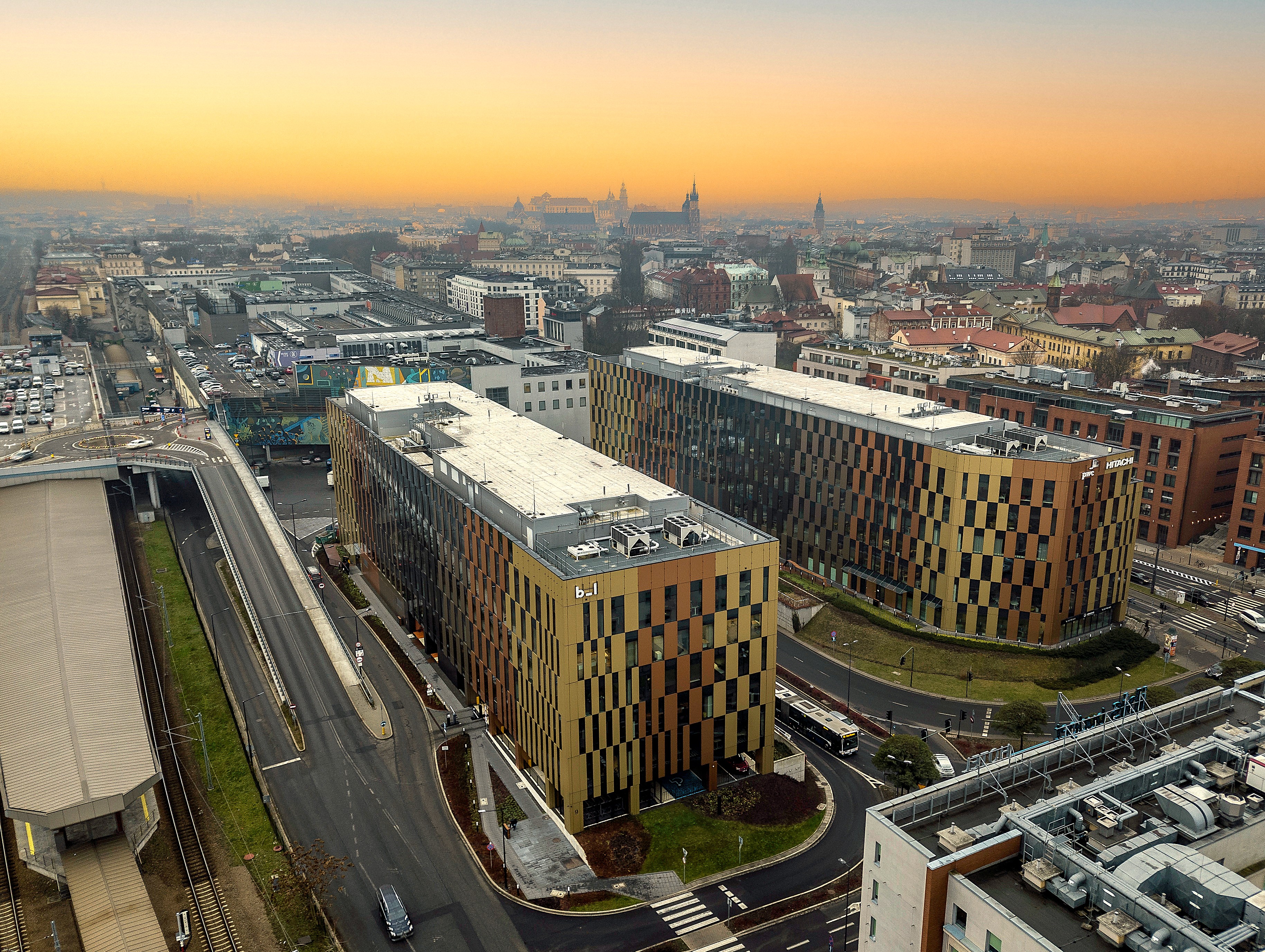 Aerial view of two modern office buildings with golden and dark vertical panel facades, located alongside curved roads. The buildings are set in a dense urban area with a sprawling cityscape in the background under a soft orange sunset sky.