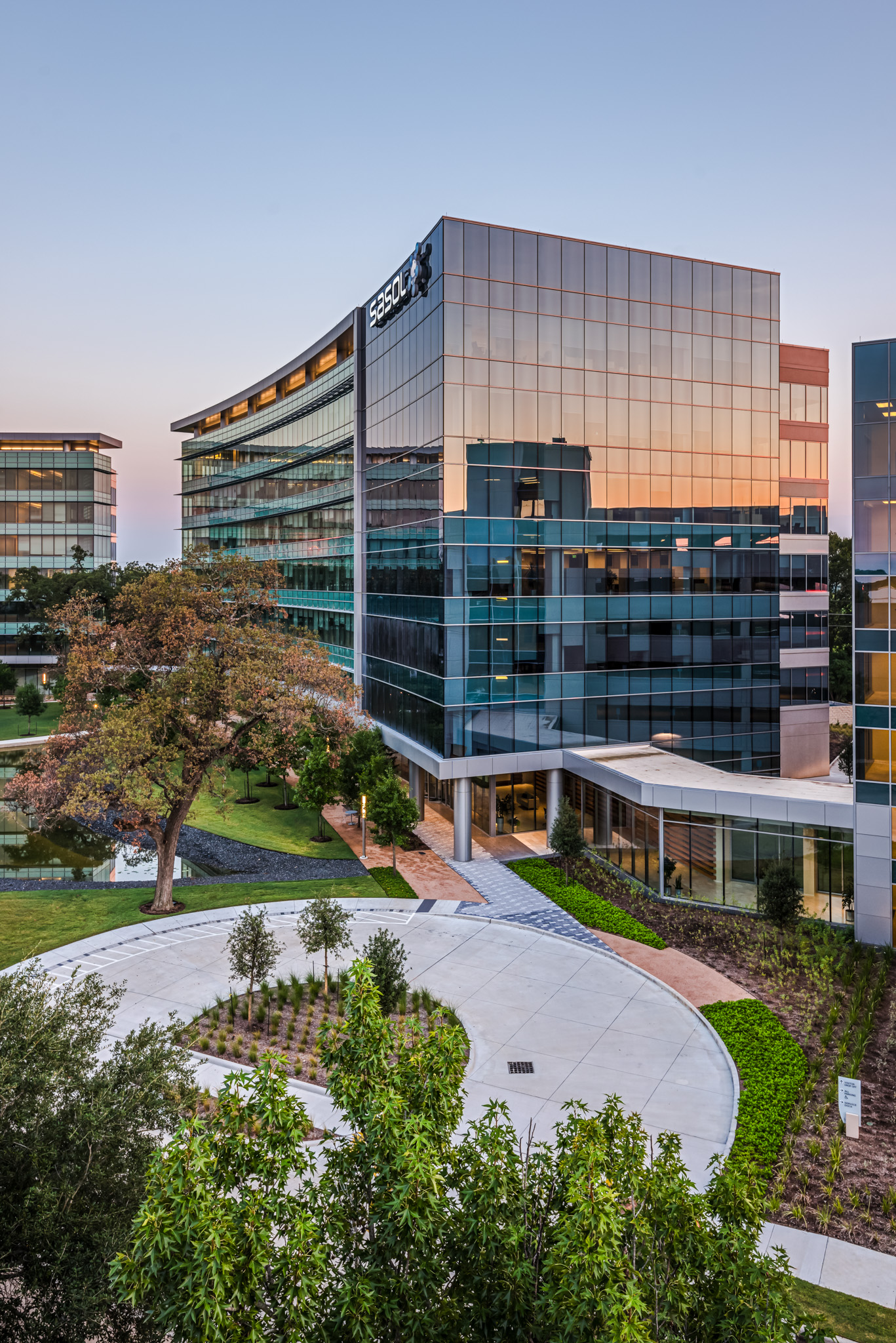 Modern office building with a glass facade reflecting the sunset, surrounded by trees and a landscaped area