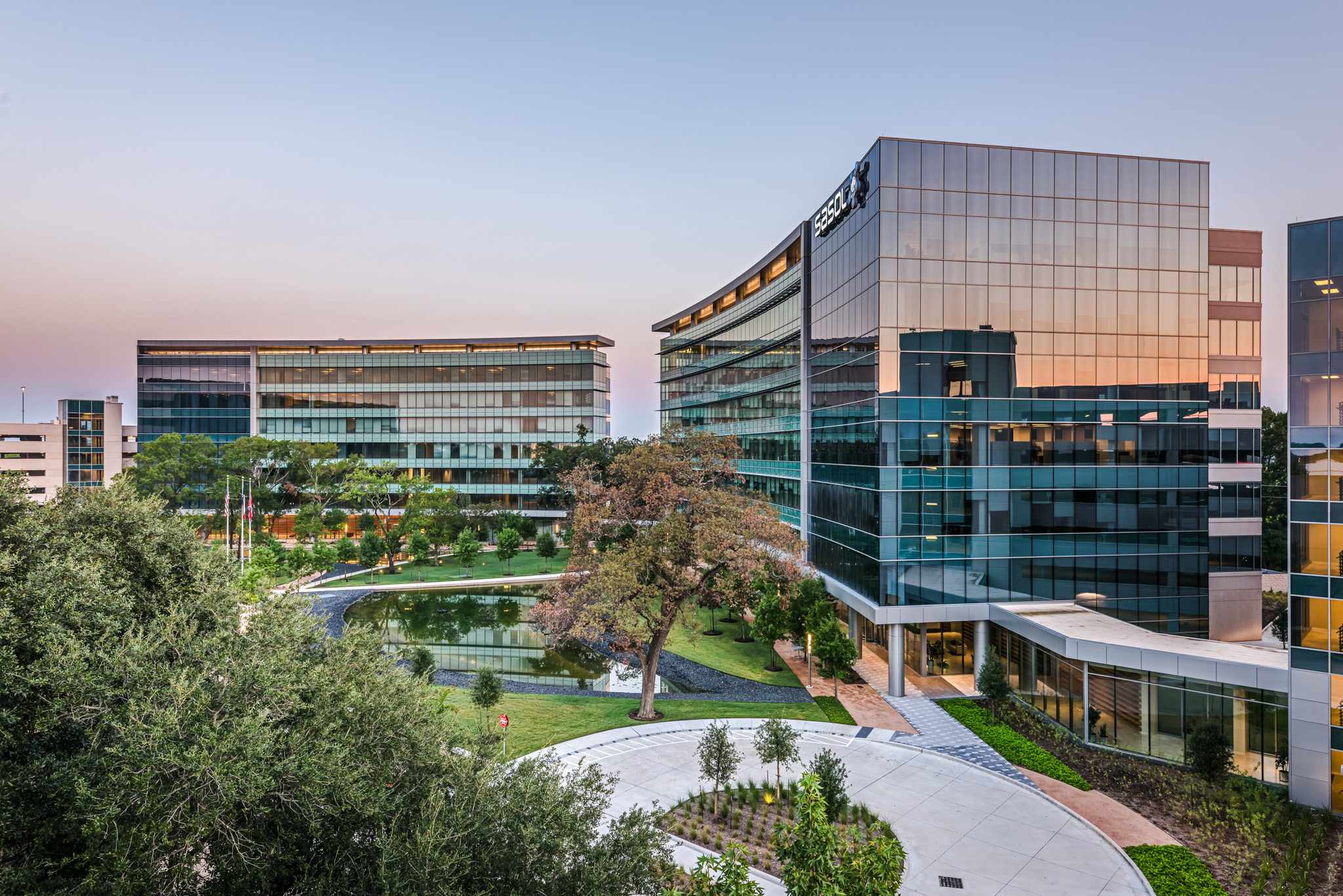 Two modern office buildings with glass facades reflecting the sunset, surrounded by a landscaped area with trees and a pond