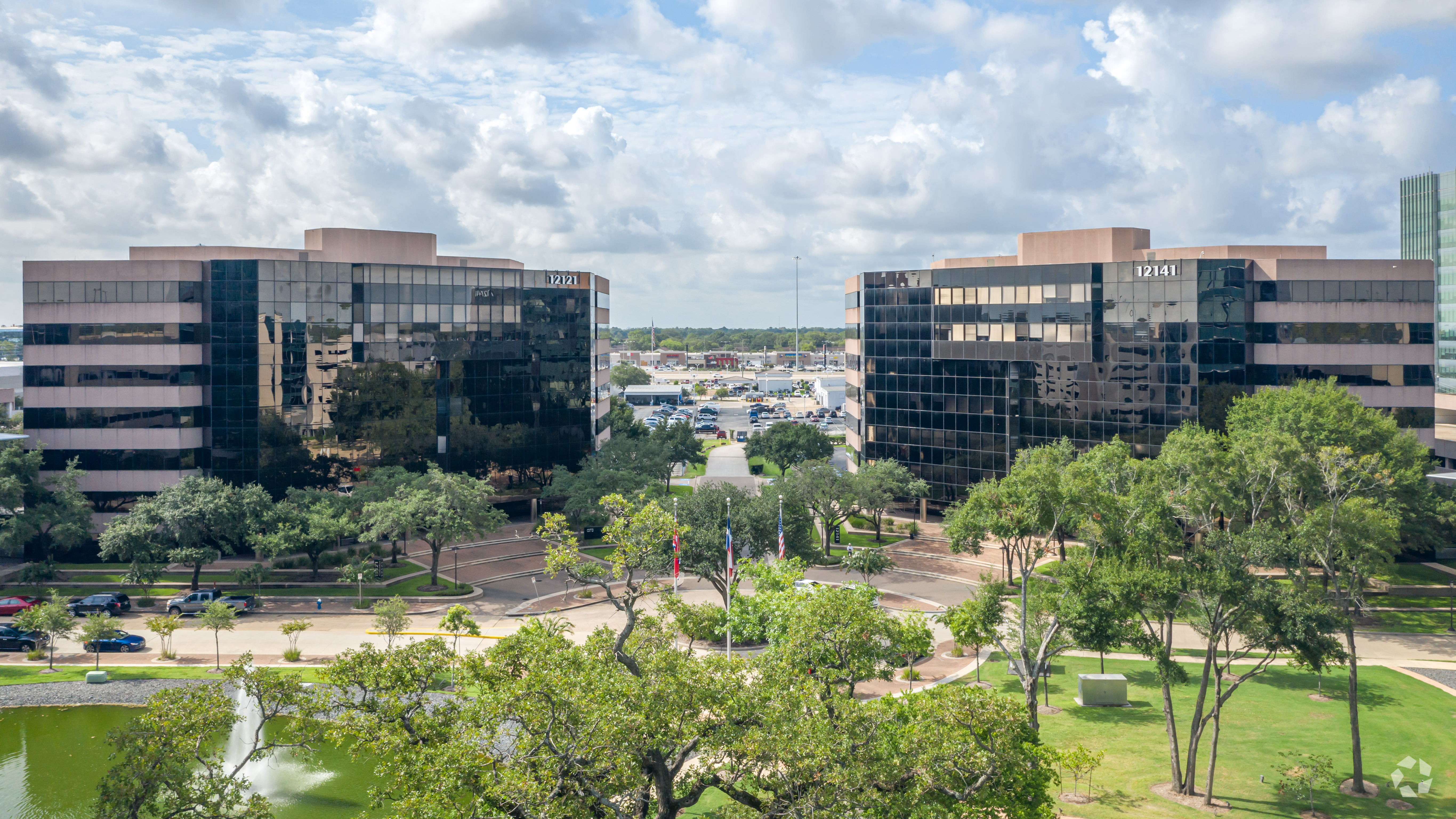 Aerial view of two identical glass office buildings marked “12141,” with a circular driveway, trees, landscaped lawns, and a fountain in the foreground.