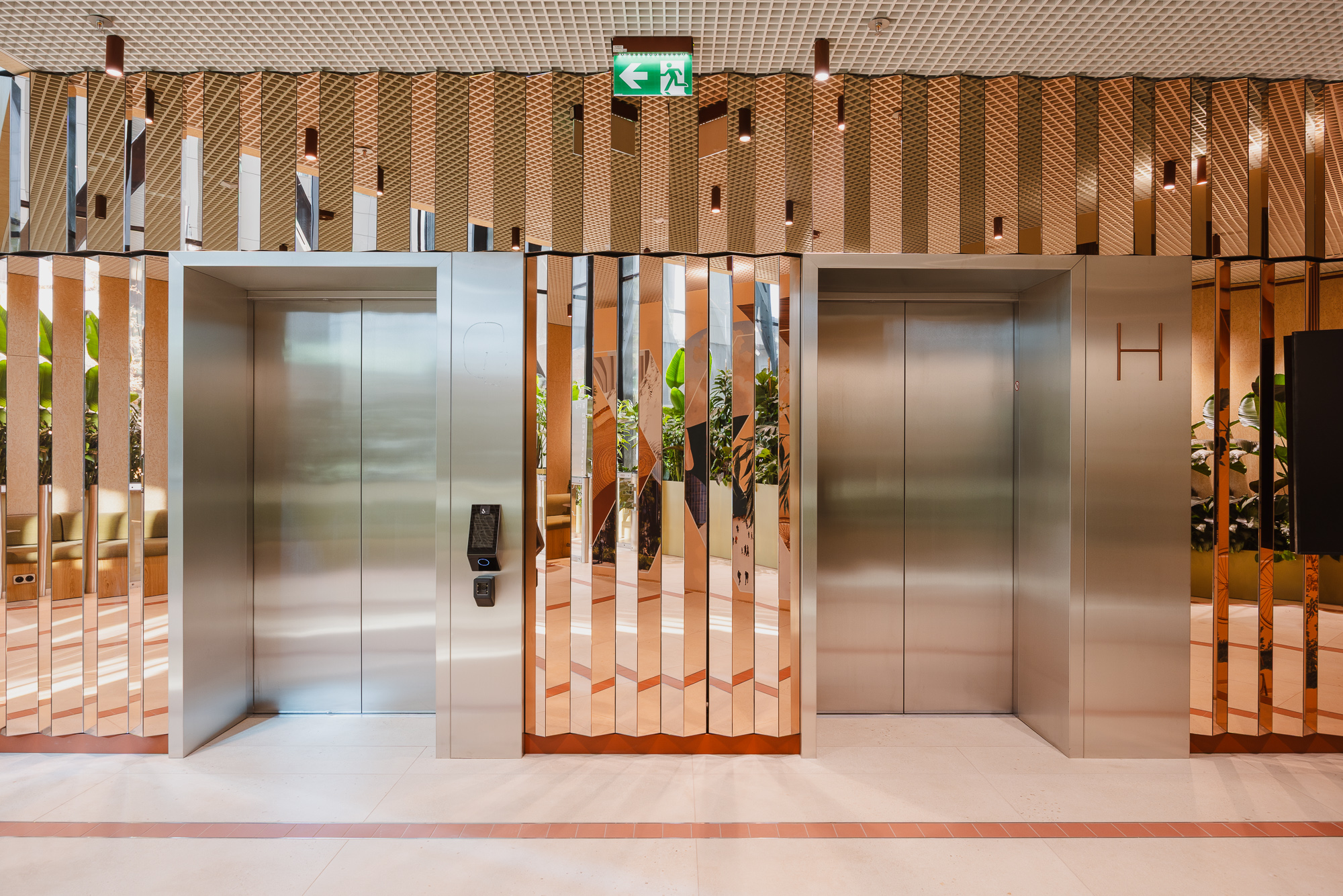  A modern elevator lobby with two stainless steel lift doors set in a wall of vertical metallic panels, warm lighting, and reflections of greenery from the adjacent interior space.
