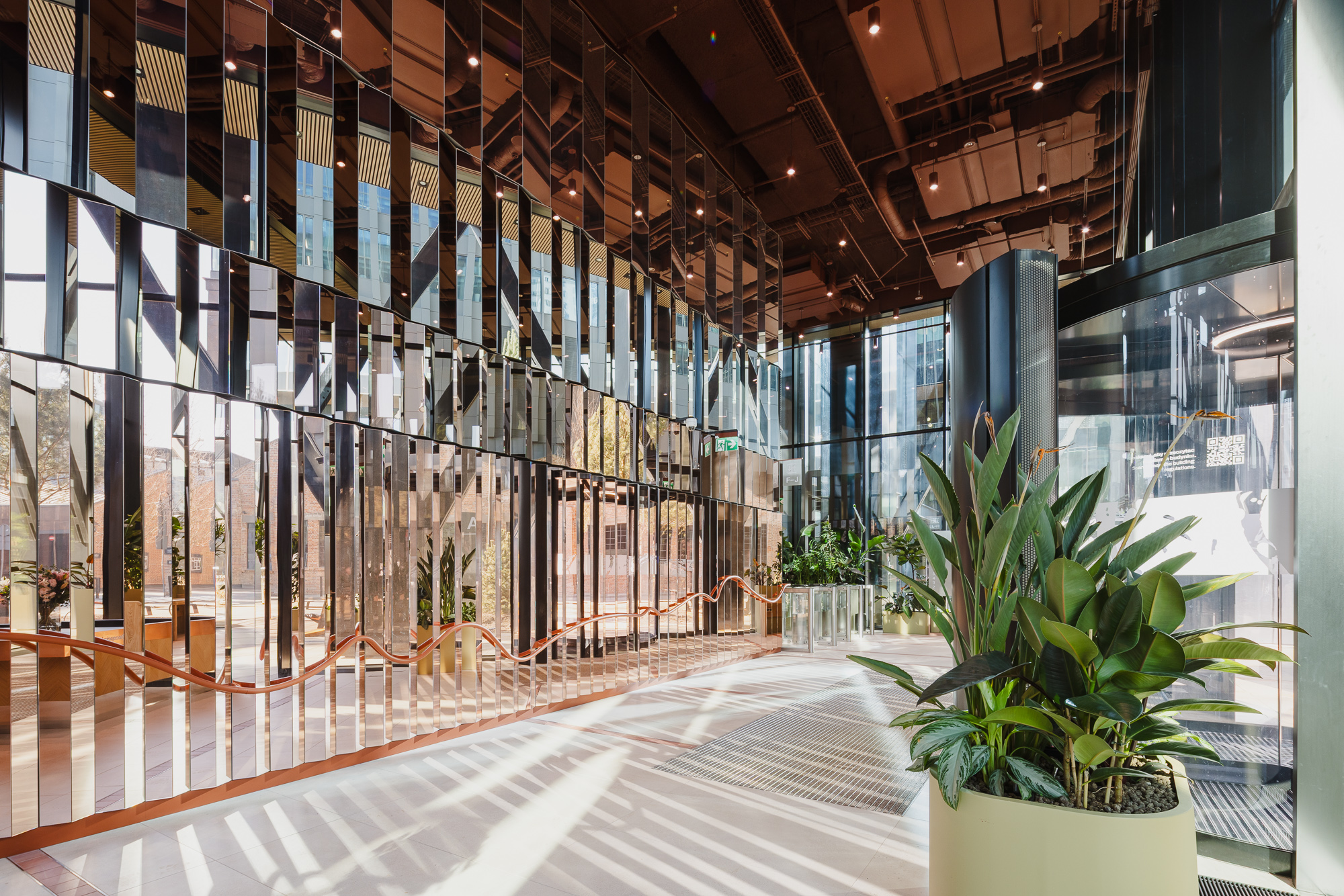 A bright, modern entrance lobby with a high ceiling, floor‑to‑ceiling glass walls, vertical mirrored metal panels, and large green plants in round pots in the foreground, with daylight streaming across the floor.