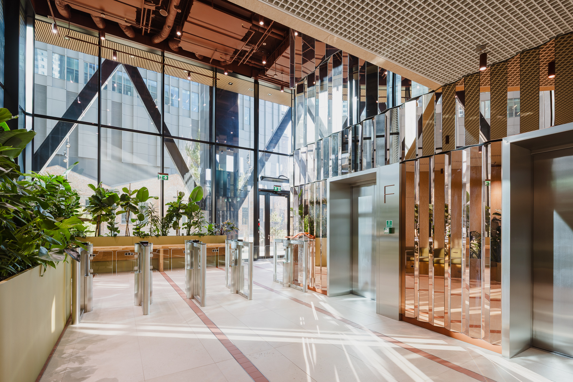 A bright, modern office lobby with floor‑to‑ceiling glass walls, metal turnstiles and elevators, polished light flooring, and abundant green plants along the sides.